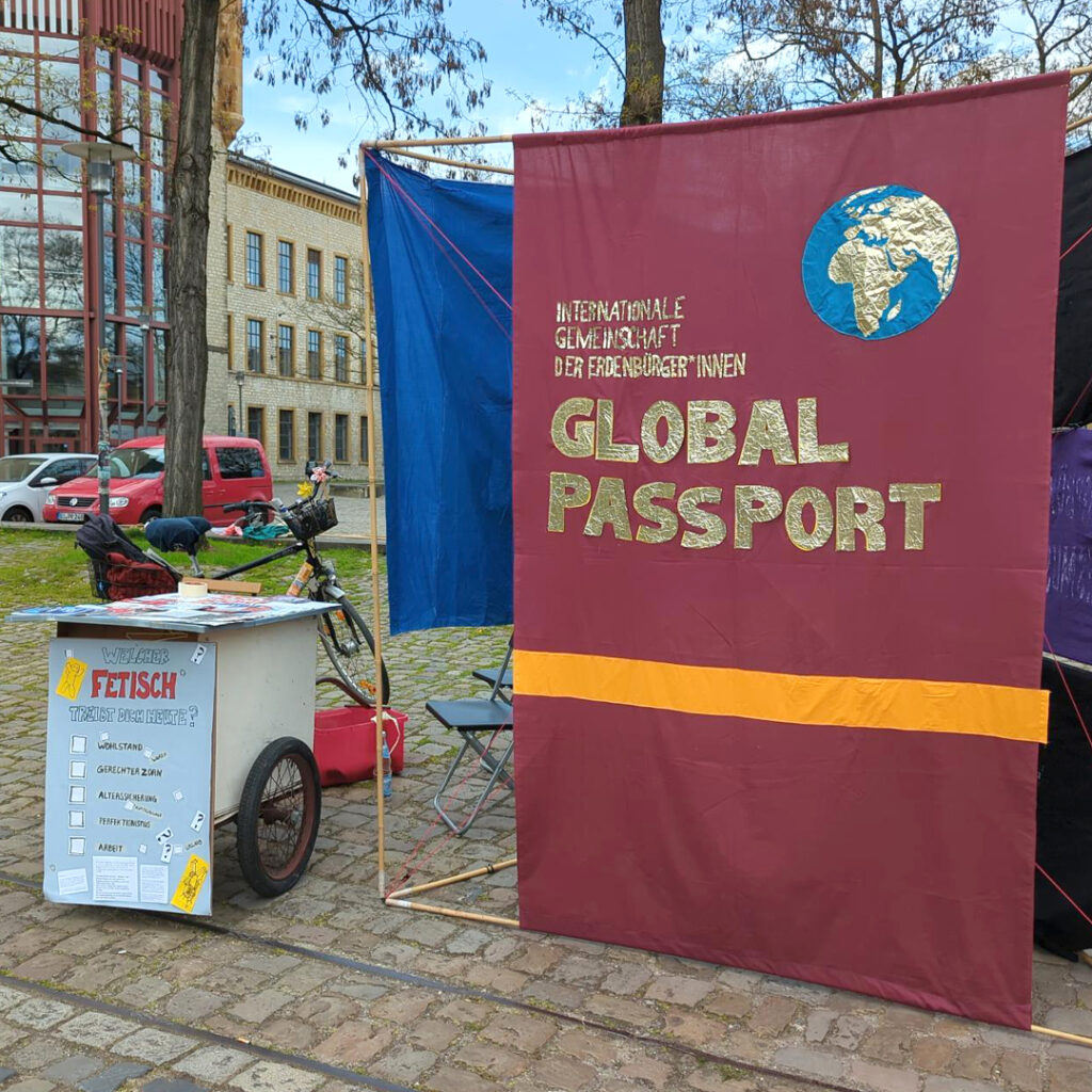Ein Bild des Infostands aus einem anderen Winkel. Ein großes Transparent, auf dem "Internationale Gemeinschaft der Erdenbürger:innen" und "Global Passport" steht, ist rechts im Bild. Es ist einem Pass nachempfunden. Links ist der Infostand mit einem Plakat davor.